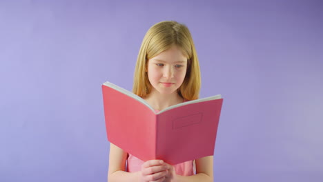 Studio-Shot-Of-Young-Girl-Studying-School-Exercise-Book-Against-Purple-Background