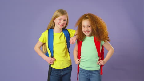 Studio-Shot-Of-Two-Girls-With-Backpacks-Going-To-School-On-Purple-Background