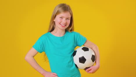 Studioaufnahme-Eines-Jungen-Mädchens,-Das-Vor-Gelbem-Hintergrund-Einen-Fußball-Unter-Dem-Arm-Hält
