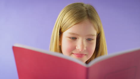 Studio-Shot-Of-Young-Girl-Studying-School-Exercise-Book-Against-Purple-Background