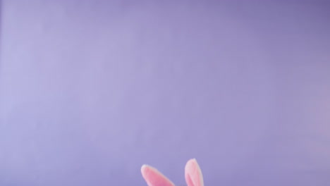 Studio-Shot-Of-Girl-Wearing-Rabbit-Ears-Jumping-Up-from-Bottom-Of-Frame-Against-Purple-Background