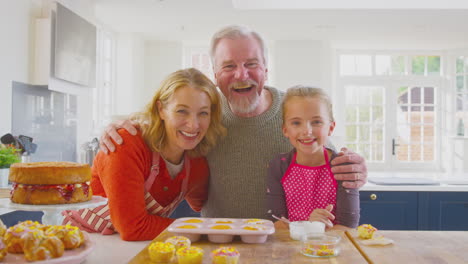 Portrait-Of-Grandparents-With-Granddaughter-Having-Fun-Decorating-Homemade-Cakes-On-Kitchen-Counter