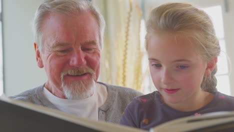 Close-Up-Of-Grandfather-And-Granddaughter-Looking-Through-Photo-Album-In-Lounge-At-Home-Together