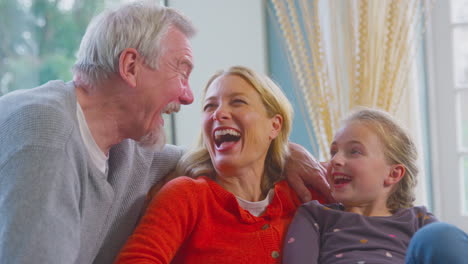 Loving-Grandparents-Hugging-Granddaughter-Sitting-On-Sofa-At-Home-Together