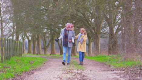 Grandfather-Gives-Granddaughter-Piggyback-Walking-Through-Winter-Countryside-With-Grandmother