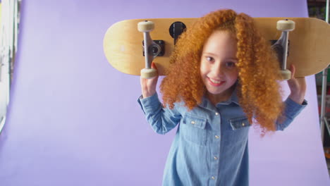 Studioaufnahme-Eines-Jungen-Mädchens,-Das-Vor-Violettem-Hintergrund-Ein-Skateboard-über-Die-Schultern-Hält