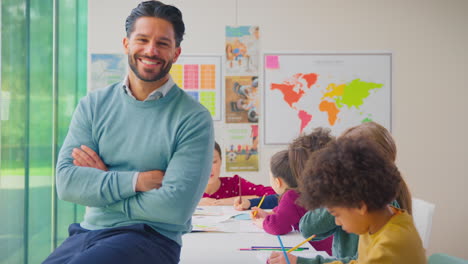 Portrait-Of-Smiling-Male-Elementary-School-Teacher-Working-At-Desk-In-Classroom-With-Students