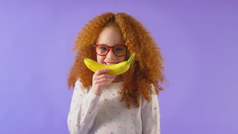 Retrato-De-Estudio-De-Una-Chica-Sosteniendo-Un-Plátano-Para-La-Boca-Sonriente-Contra-El-Fondo-Púrpura