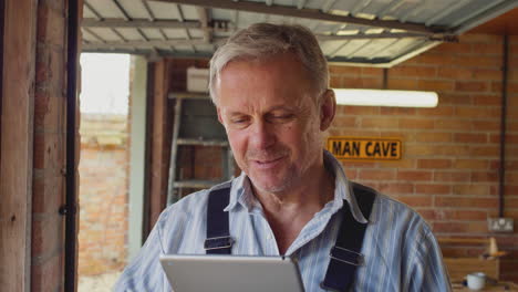 Mature-Male-Wearing-Overalls-Using-Digital-Tablet-In-Garage-Workshop