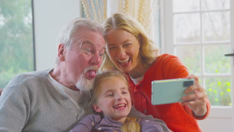 Abuelos-Con-Nieta-Haciendo-Muecas-Posando-Juntos-Para-Un-Selfie-En-Un-Teléfono-Móvil-En-Casa