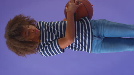 Vertikales-Video-Eines-Lächelnden-Jungen,-Der-Vor-Violettem-Hintergrund-Tricks-Mit-Basketball-Macht