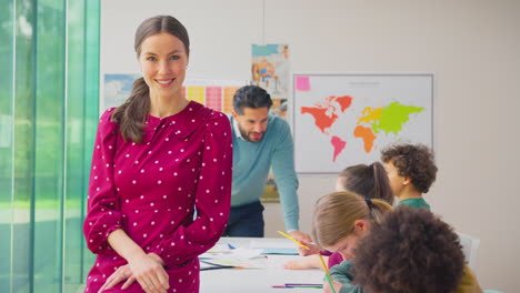 Portrait-Of-Smiling-Female-Elementary-School-Teacher-Working-At-Desk-In-Classroom-With-Students