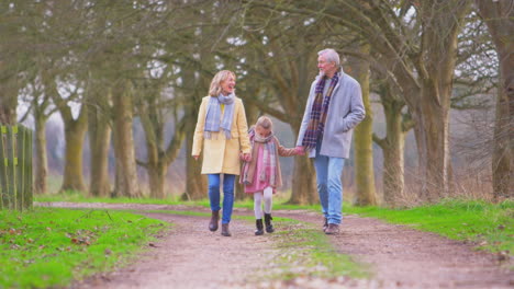 Grandparents-With-Granddaughter-Outside-Walking-Through-Winter-Countryside-Holding-Hands