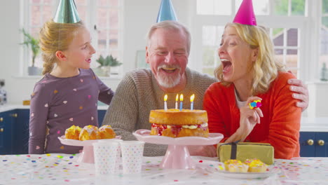 Grandparents-With-Granddaughter-Celebrating-Blowing-Out-Candles-On-Birthday-Cake-At-Home-Together