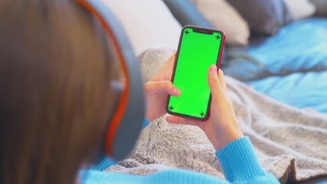 Teenagermädchen-Auf-Dem-Sofa-Mit-Kabellosen-Kopfhörern,-Das-Inhalte-Von-Einem-Greenscreen-Handy-Streamt