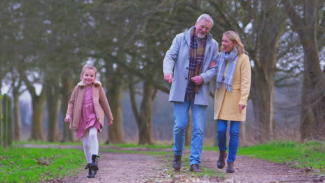 Grandparents-Watching-Granddaughter-Skipping-Ahead-On-Outside-Walking-Through-Winter-Countryside