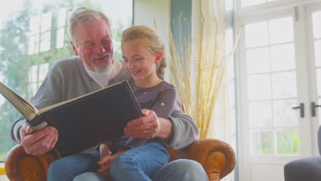 Grandfather-And-Granddaughter-Looking-Through-Photo-Album-In-Lounge-At-Home-Together