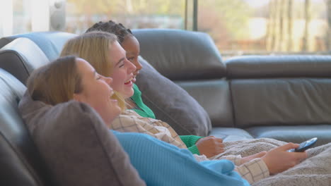 Group-Of-Multi-Cultural-Teenage-Girl-Friends-Snuggled-Under-Blanket-Watching-TV-At-Home