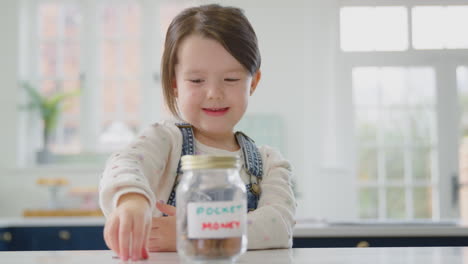 Girl-Saving-Money-Into-Jar-Labelled-Pocket-Money-On-Kitchen-Counter-At-Home