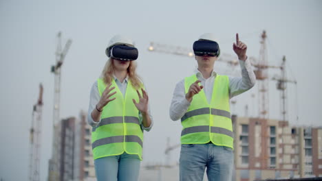 Two-people-Man-and-woman-wearing-virtual-reality-glasses-on-the-background-of-buildings-under-construction-with-cranes.-Manager-and-assistant-on-design-with-your-hands-mimicking-the-interface-of-the-application