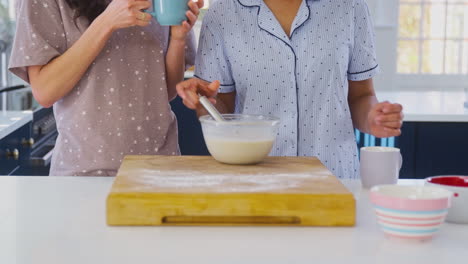 Same-Sex-Female-Couple-In-Pyjamas-Making-Morning-Pancakes-And-Drinking-Coffee-In-Kitchen-At-Home
