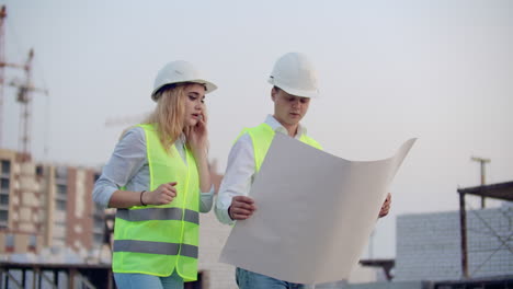 Workers-with-drawings-at-the-construction-site.-Two-workers-man-and-woman-in-protective-harhats-working-with-drawings-at-the-construction-site-outdoors.