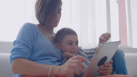 Happy-family-mom-and-kid-son-using-digital-tablet-sitting-on-sofa-smiling-parent-mother-with-child-son-holding-pc-computer-looking-at-screen-do-online-shopping-make-video-call-watch-cartoons