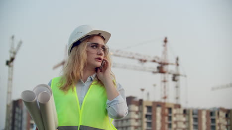 Talking-woman-in-a-helmet-on-the-phone-on-the-background-of-construction-with-cranes-holding-drawings-in-hand.-Female-engineer-on-construction-site