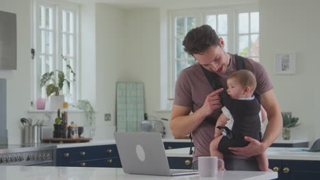 Transgender-Father-Working-From-Home-On-Laptop-Looking-After-Baby-Son-In-Sling