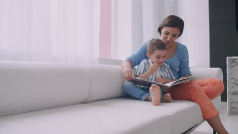 Liebevolle-Mutter-Lehrt-Ihren-Sohn-Zu-Lesen-Halten-Buch-Sprechen-Auf-Der-Couch-Zu-Hause