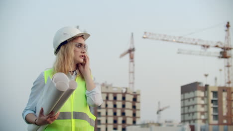 Talking-woman-in-a-helmet-on-the-phone-on-the-background-of-construction-with-cranes-holding-drawings-in-hand.-Female-engineer-on-construction-site