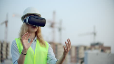 A-woman-in-virtual-reality-glasses-helmet-and-vest-on-the-background-of-construction-controls-the-hands-of-the-interface-and-checks-the-quality-of-construction-and-development-of-the-project-and-the-development-plan-and-landscape.-Landscape-designer-uses-virtual-reality