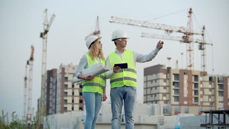 Two-colleagues-of-engineers-a-man-and-a-woman-discussing-a-drawing-and-a-tablet-computer-on-the-background-of-buildings-under-construction-and-cranes-a-woman-talking-on-the-phone-with-the-boss