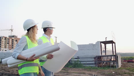 The-builder-and-architect-man-and-woman-are-discussing-the-construction-plan-of-the-modern-business-center-standing-near-construction-site.-They-go-to-the-building.