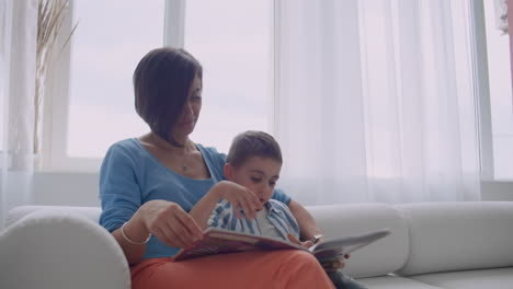 Liebevolle-Mutter-Lehrt-Ihren-Sohn-Zu-Lesen-Halten-Buch-Sprechen-Auf-Der-Couch-Zu-Hause