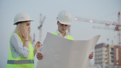 Two-builders-with-drawings-standing-on-the-background-of-buildings-under-construction-in-helmets-and-vests-a-woman-talking-on-the-phone-with-the-customer.