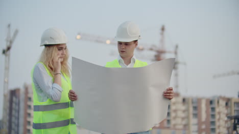 Two-colleagues-engineers-man-and-woman-discussing-the-drawing-on-the-background-of-buildings-and-cranes-a-woman-talking-on-the-phone-with-the-boss