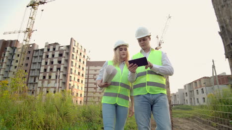 Business-building-industry-technology-and-people-concept---smiling-builder-in-hardhat-with-tablet-pc-computer-along-with-woman-with-drawings-of-builders-at-construction-site