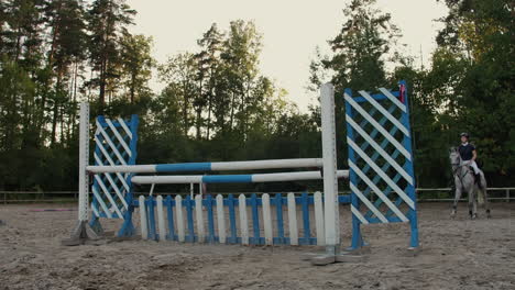 SLOW-MOTION-CLOSE-UP-LOW-ANGLE:-Horsegirl-riding-strong-brown-horse-jumping-the-fence-in-sunny-outdoors-sandy-parkour-dressage-arena.-Competitive-rider-training-jumping-over-obstacles-in-manege