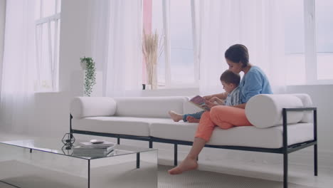 A-young-mother-with-a-child-reading-a-book-sitting-in-a-bright-white-interior-of-the-house-in-the-living-room-on-the-couch