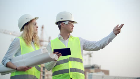 Two-colleagues-of-engineers-a-man-and-a-woman-discussing-a-drawing-and-a-tablet-computer-on-the-background-of-buildings-under-construction-and-cranes-a-woman-talking-on-the-phone-with-the-boss