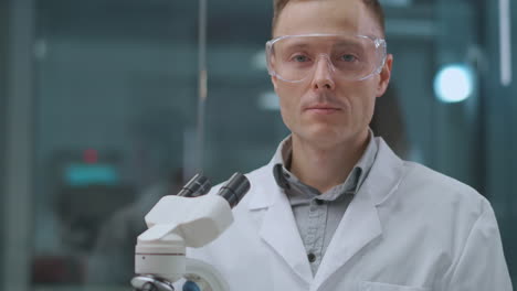 portrait-of-confident-laboratory-technician-man-in-working-room-wearing-protective-glasses-standing-near-microscope-chemist-or-health-professional
