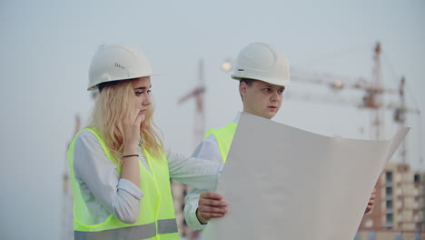 Two-builders-with-drawings-standing-on-the-background-of-buildings-under-construction-in-helmets-and-vests-a-woman-talking-on-the-phone-with-the-customer.