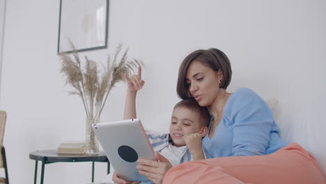 Mom-and-son-looking-at-the-tablet-screen-lying-on-a-white-bed.-Play-games-with-your-son-on-your-tablet-computer-and-watch-funny-videos.
