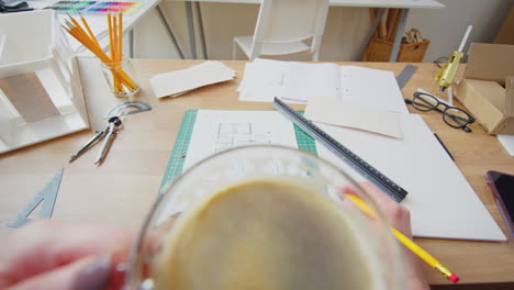 POV-Shot-Of-Female-Architect-Drinking-Cup-Of-Coffee-Working-In-Office-On-Plans-For-New-Building