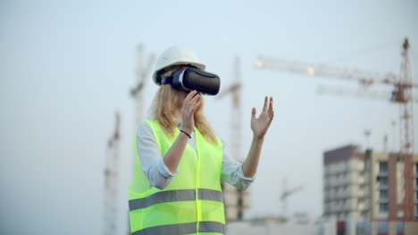 A-woman-in-virtual-reality-glasses-helmet-and-vest-on-the-background-of-construction-controls-the-hands-of-the-interface-and-checks-the-quality-of-construction-and-development-of-the-project-and-the-development-plan-and-landscape.-Landscape-designer-uses-virtual-reality