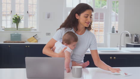 Mutter-Mit-Kleinem-Sohn-Arbeitet-Von-Zu-Hause-Aus-Am-Laptop-In-Der-Küche-Und-Reinigt-Die-Oberfläche