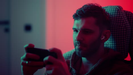 addicted-gamer-playing-by-smartphone-new-app-with-video-games-portrait-of-emotional-young-man-at-home-in-night-listening-to-sound-by-in-ear-headphones