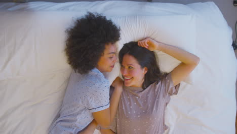 Overhead-Shot-Of-Loving-Same-Sex-Female-Couple-Wearing-Pyjamas-Lying-On-Bed-At-Home
