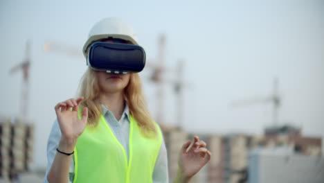 Woman-designer-on-a-building-site-in-hard-hat-and-vest-in-the-glasses-of-virtual-reality-to-move-your-hands-mimicking-the-interface-on-the-background-of-the-cranes-at-sunset.